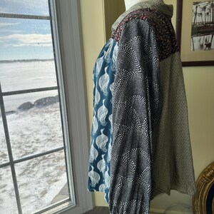 May include: A long-sleeved shirt with a patchwork design. The shirt is made of different fabrics with various patterns, including a teal fabric with white bird silhouettes, a black fabric with white polka dots, and a brown fabric with a subtle pattern.
