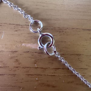 May include: Close-up of a silver necklace clasp and chain. The chain is delicate, with small, interconnected links. The clasp is a spring ring type, designed for secure fastening. The background is a light brown wooden surface.