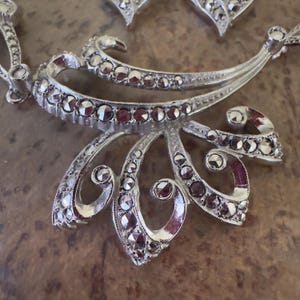 May include: Silver-toned necklace and earring set with a floral design. The jewelry features a series of small, round, clear stones and a dark red-brown accent. The necklace has a central, ornate pendant with curved and leaf-like elements.