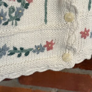 May include: White knitted cardigan with a scalloped hem and floral embroidery in shades of pink, blue, and green. The cardigan has two buttons.