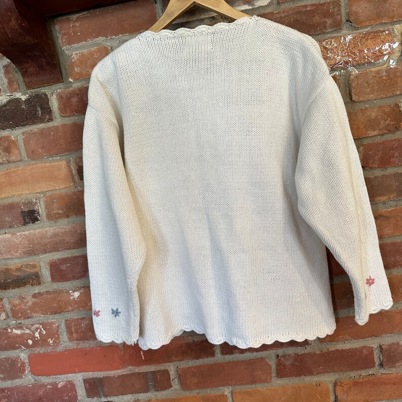 May include: A white knitted cardigan with a scalloped hem and embroidered flowers on the cuffs. The cardigan is made of a soft, lightweight material and has a relaxed fit.