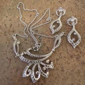 May include: A silver-toned jewelry set featuring a necklace and matching earrings. The necklace has a floral design with a pendant and chain. The earrings are teardrop-shaped. The set is embellished with small, sparkling stones.