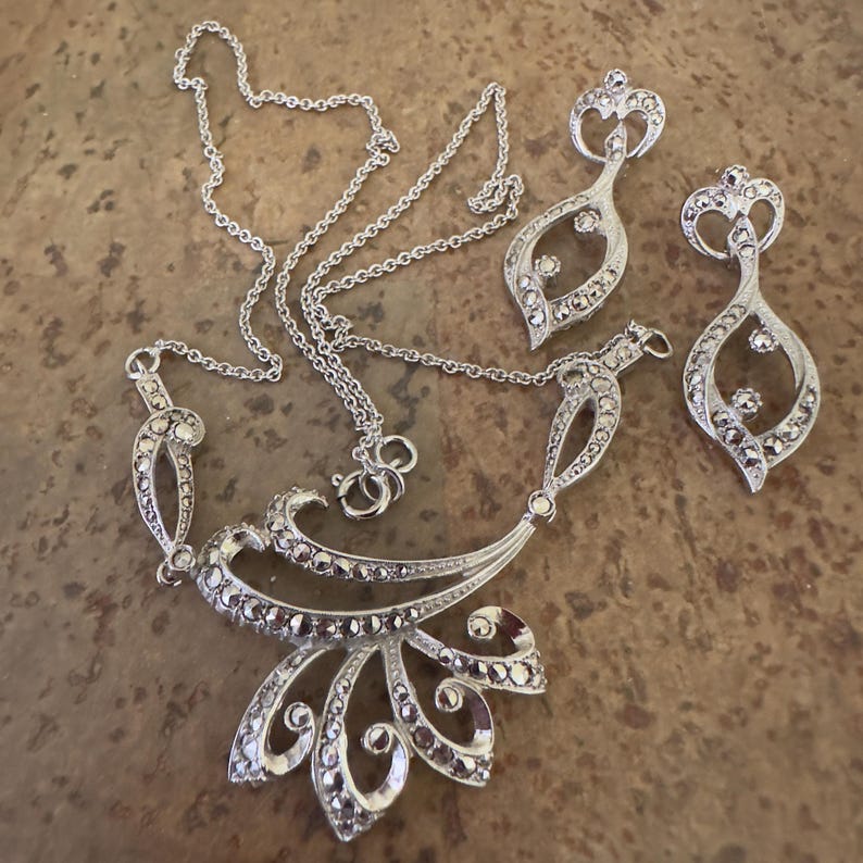 May include: A silver-toned jewelry set featuring a necklace and matching earrings. The necklace has a floral design with a pendant and chain. The earrings are teardrop-shaped. The set is embellished with small, sparkling stones.