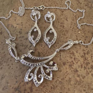May include: A silver-toned jewelry set featuring a necklace and matching earrings. The necklace has a floral design with a pendant, and the earrings are teardrop-shaped. The set is adorned with small, sparkling stones.