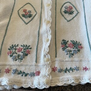 May include: A cream knitted cardigan with a scalloped hem and floral embroidery. The cardigan has a button-up front and green stripes.