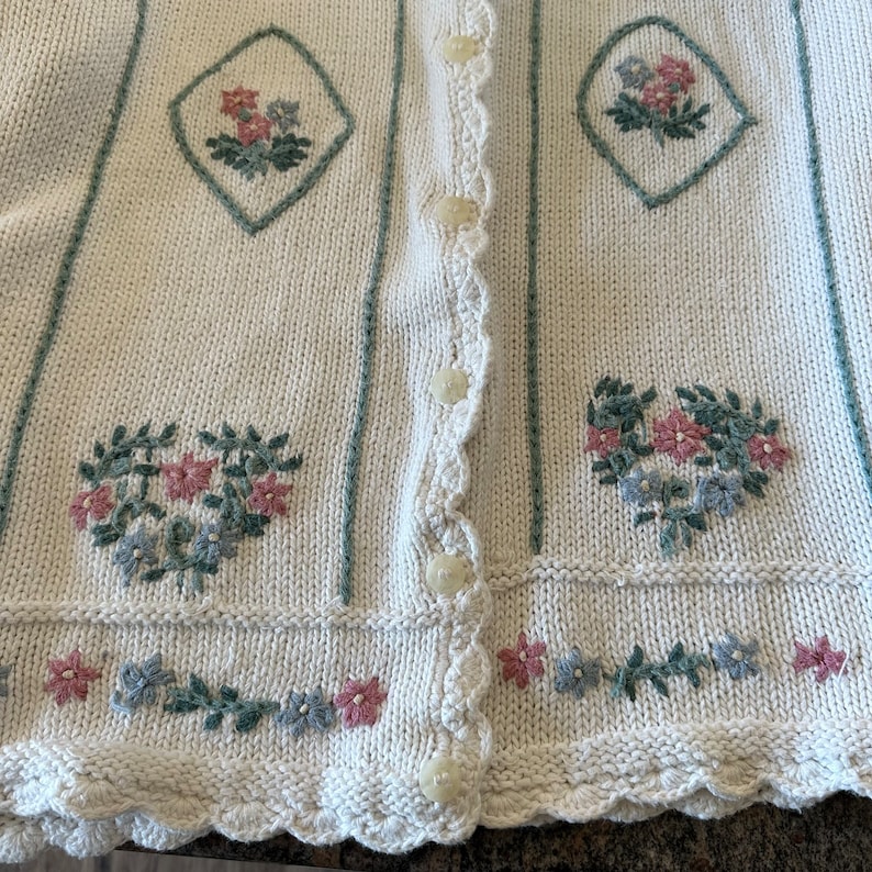 May include: A cream knitted cardigan with a scalloped hem and floral embroidery. The cardigan has a button-up front and green stripes.
