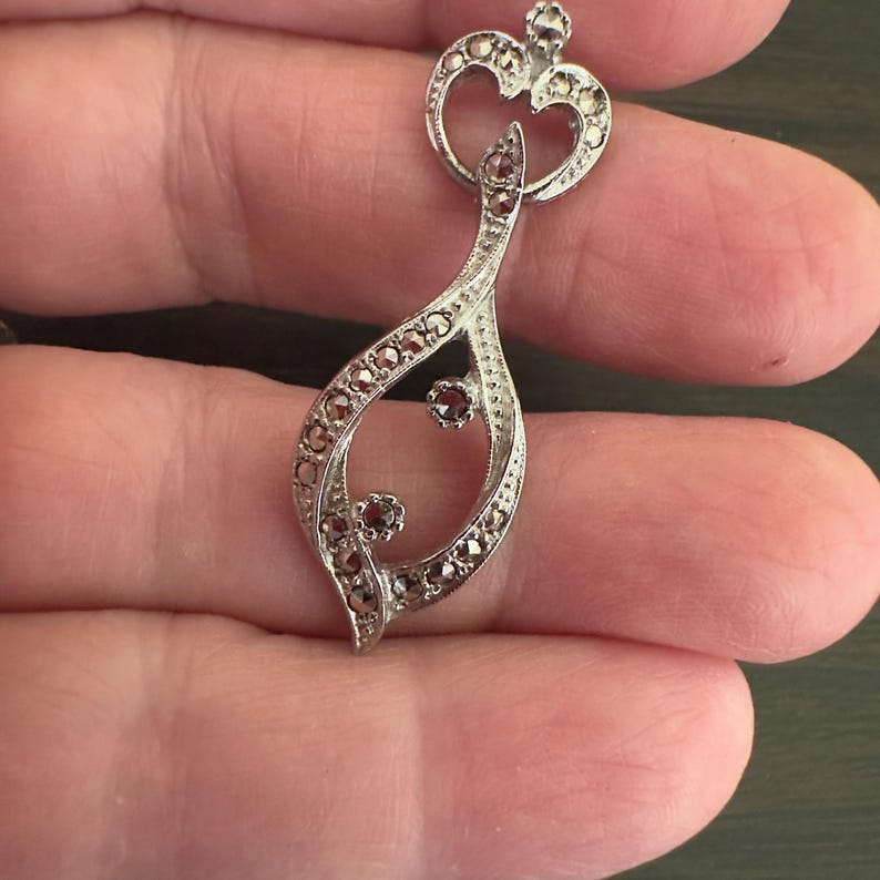 May include: Silver-toned pendant with a teardrop design, adorned with small, dark red gemstones and tiny silver accents. The pendant features a heart-shaped top and a delicate, openwork pattern.