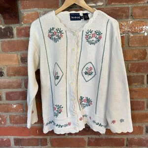 May include: A white cardigan with a scalloped hem and floral embroidery. The cardigan has a button closure and is made by Weekend Edition.