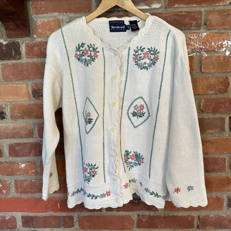 May include: A white cardigan with a scalloped hem and floral embroidery. The cardigan has a button closure and is made by Weekend Edition.