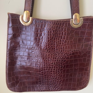 May include: A brown faux crocodile-skin tote bag with a rectangular shape. The bag has two handles and gold-colored metal accents. The bag's surface has a textured, embossed pattern.