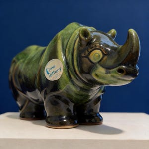 May include: A ceramic rhino figurine in shades of green and brown, with a glossy finish. The rhino has a prominent horn and is labeled with a sticker that reads "Dine Pottery". The figurine is displayed on a white surface against a blue backdrop.