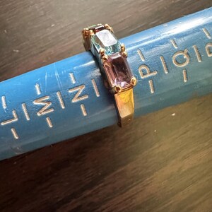 K&ouml;nnte beinhalten: Ein goldener Ring mit drei rechteckigen Edelsteinen. Die Edelsteine sind blau, hellblau und lila.