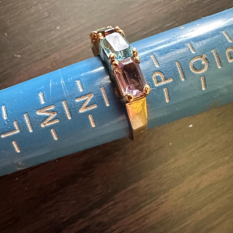 K&ouml;nnte beinhalten: Ein goldener Ring mit drei rechteckigen Edelsteinen. Die Edelsteine sind blau, hellblau und lila.
