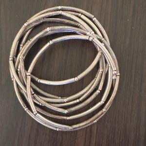 May include: A collection of silver-coloured bracelets. Each bracelet is made of curved, tubular segments connected by small, round beads. The bracelets are arranged in a circular pattern, creating a layered effect. The metal has a slightly aged appearance.