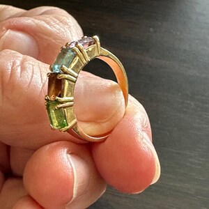 K&ouml;nnte beinhalten: Ein Goldring mit f&uuml;nf rechteckigen Edelsteinen. Die Edelsteine sind gr&uuml;n, braun, blau und lila.