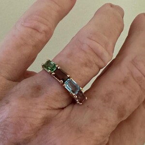 K&ouml;nnte beinhalten: Ein goldener Ring mit einer Reihe von smaragdgr&uuml;nen, burgunderroten und hellblauen Edelsteinen.