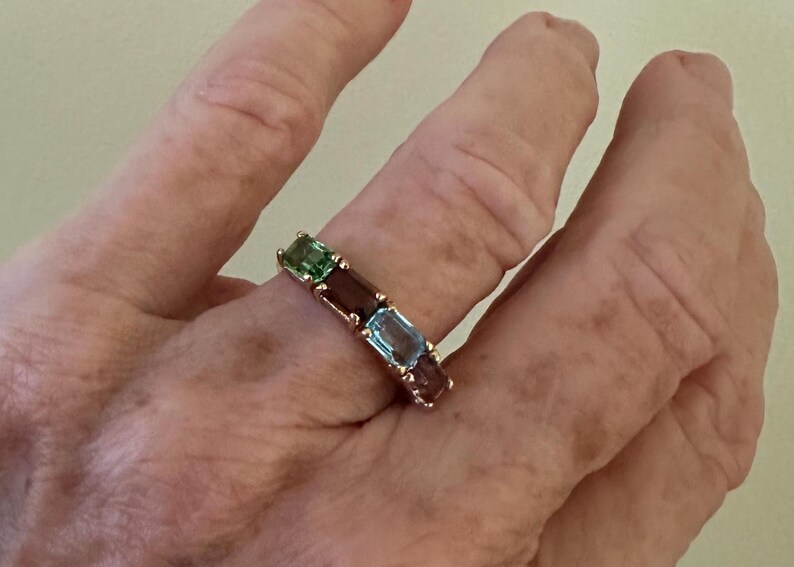 K&ouml;nnte beinhalten: Ein goldener Ring mit einer Reihe von smaragdgr&uuml;nen, burgunderroten und hellblauen Edelsteinen.