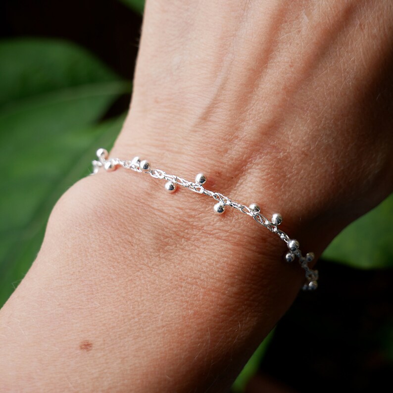 Pode incluir: Um bracelete de corrente de prata com pequenas contas de prata penduradas.