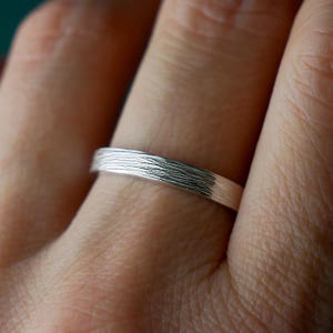 Puede incluir: Un anillo de banda de plata con una textura horizontal fina y distintiva que imita la apariencia de la veta de la madera natural. La superficie metálica tiene un acabado pulido con líneas oscuras hundidas que resaltan el patrón lineal.