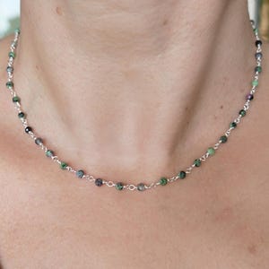Può includere: Una delicata collana con piccole perline rotonde nei toni del verde, nero e viola infilate su una catena d'argento. La collana è esposta attorno al collo di una persona, mostrando la sua sottile eleganza e il suo design.