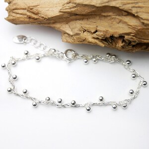 Pode incluir: Um bracelete de corrente de prata com pequenas contas de prata penduradas nele. O bracelete tem um fecho de lagosta.