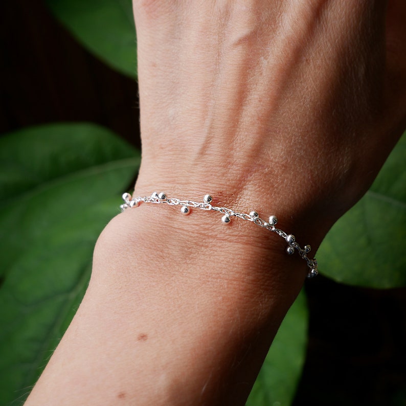 Pode incluir: Um bracelete de corrente de prata com pequenas contas de prata penduradas.