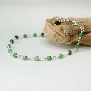 May include: A silver bracelet with small, round, green beads. The bracelet is displayed on a piece of driftwood. The beads are evenly spaced along the silver chain. The clasp is visible, adding to the overall aesthetic of the jewelry.