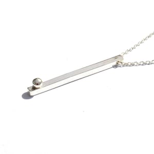 Op de afbeelding: Een zilveren ketting met een lange, rechthoekige hanger en een kleine, ronde kraal aan de bovenkant.