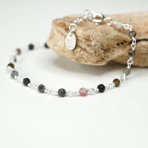 May include: A delicate bracelet with a silver chain, adorned with a series of multi-coloured, round gemstones. The gemstones include pink, green, black, and grey hues. The bracelet is displayed on a piece of wood.