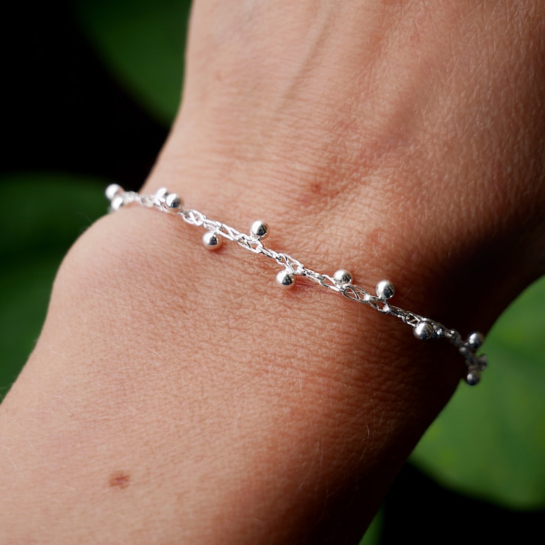 Pode incluir: Um bracelete de corrente de prata com pequenas contas de prata penduradas.