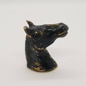 May include: A black horse head figurine with gold detailing. The horse head is facing right, with a red gem eye. The figurine has a distressed, vintage appearance.