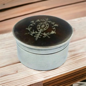 May include: A small, round, silver-coloured container with a dark brown lid. The lid features an ornate, silver-coloured floral design. The container sits on a light brown wooden surface.