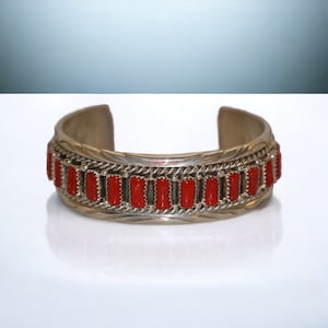 May include: Silver cuff bracelet with a row of vibrant red rectangular stones. The bracelet has a detailed, engraved design around the stones and along the edges. A classic piece of jewelry.