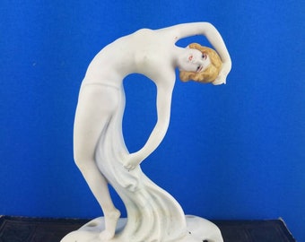 May include: A white porcelain figurine of a woman in a flowing gown, bending backward. The figurine has blonde hair and is mounted on a base with several holes. The background is a solid blue.