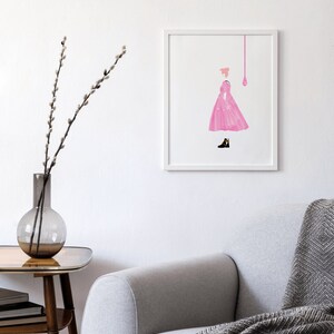 Puede incluir: Una impresi&oacute;n art&iacute;stica enmarcada con una ilustraci&oacute;n rosa de una persona con un vestido, con una forma de l&aacute;grima rosa encima. La obra de arte est&aacute; sobre un fondo blanco. Un sof&aacute; gris con una manta estampada y una mesa auxiliar con un jarr&oacute;n est&aacute;n en primer plano.