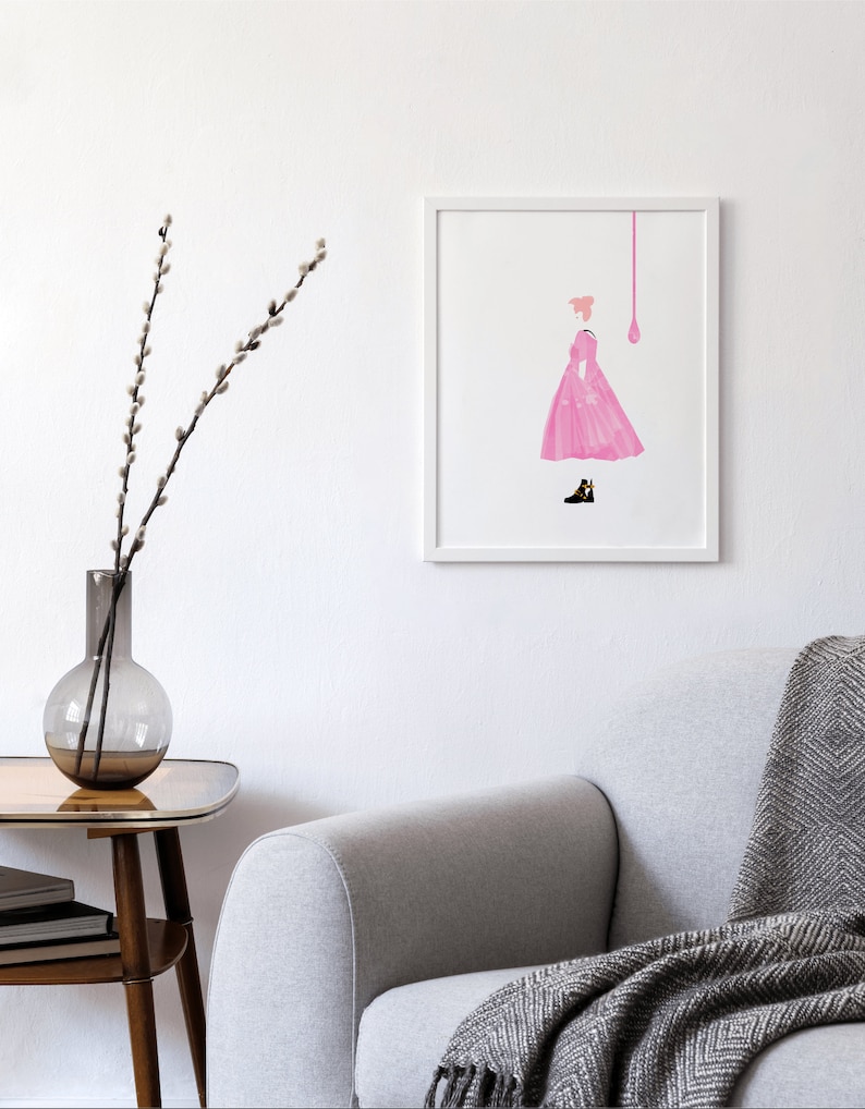 Puede incluir: Una impresi&oacute;n art&iacute;stica enmarcada con una ilustraci&oacute;n rosa de una persona con un vestido, con una forma de l&aacute;grima rosa encima. La obra de arte est&aacute; sobre un fondo blanco. Un sof&aacute; gris con una manta estampada y una mesa auxiliar con un jarr&oacute;n est&aacute;n en primer plano.