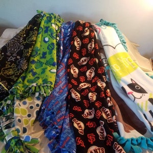 May include: A collection of fleece blankets with various patterns and colors. Designs include a black blanket with white and red robot figures, a green blanket with blue and green shapes, a blue blanket with white text, and a black blanket with the San Francisco Giants logo.
