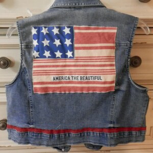 May include: A denim vest with a patriotic design. The back features a patch of the American flag with the text "AMERICA THE BEAUTIFUL." A red stripe accents the bottom hem. The vest is sleeveless and has a classic collar.