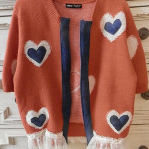May include: An orange cardigan sweater with white heart-shaped designs and navy blue hearts. The sweater has a denim-like trim and white lace detailing at the bottom. The brand name "SHEIN" is visible on the label.