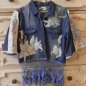 May include: A denim shirt with white floral designs and lace accents. The shirt features short sleeves with patterned fabric and lace trim. The bottom hem has a blue lace fringe.