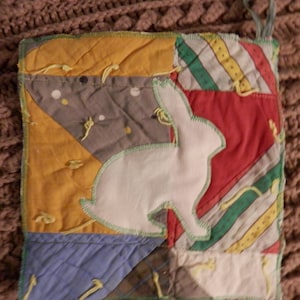 May include: A patchwork pot holder with a white bunny applique. The pot holder features a variety of fabrics in yellow, gray, red, and blue, with green stitching and a hanging loop. The design is a mix of geometric shapes and stripes.