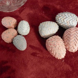 May include: A collection of decorative, egg-shaped objects in various colors, including pink, white, and gray. The objects have a woven or textured appearance, and are arranged on a deep red surface. The objects are likely for home decor.