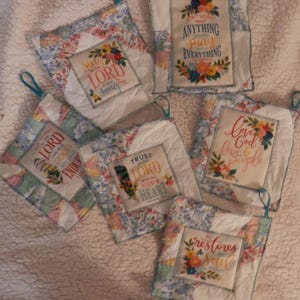 May include: A collection of quilted pot holders with floral patterns and inspirational quotes. Each square pot holder features a different message, such as "Sing to the Lord a new song" and "Trust the Lord with all your heart."