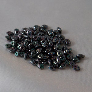 May include: A collection of dark, iridescent beads, each with two holes. The beads have a glossy finish, reflecting green, blue, and purple hues. They are scattered on a light grey, textured surface.