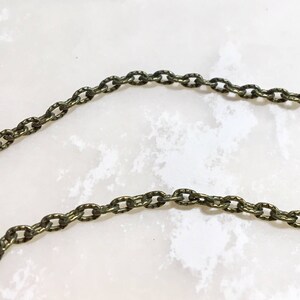 May include: Close-up of a bronze-coloured chain with oval links. The chain is set against a white and grey marbled background. The chain is likely used for jewellery or other decorative purposes.
