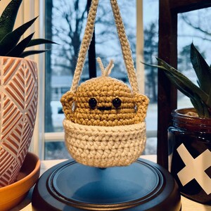 May include: A handmade crochet hanging planter in beige and brown. The planter features a small, smiling face and is suspended by a braided beige cord. The planter is designed to hold small plants and adds a touch of whimsy to any space.