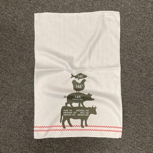May include: White kitchen towel with a red and white striped border. The towel features a graphic of a fish, chicken, pig, and cow with corresponding internal cooking temperatures in Fahrenheit.