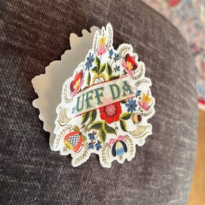 diecut adhesive sticker features colorful Norwegian rosemaling with the phrase ‘Uff Da’ written across the center