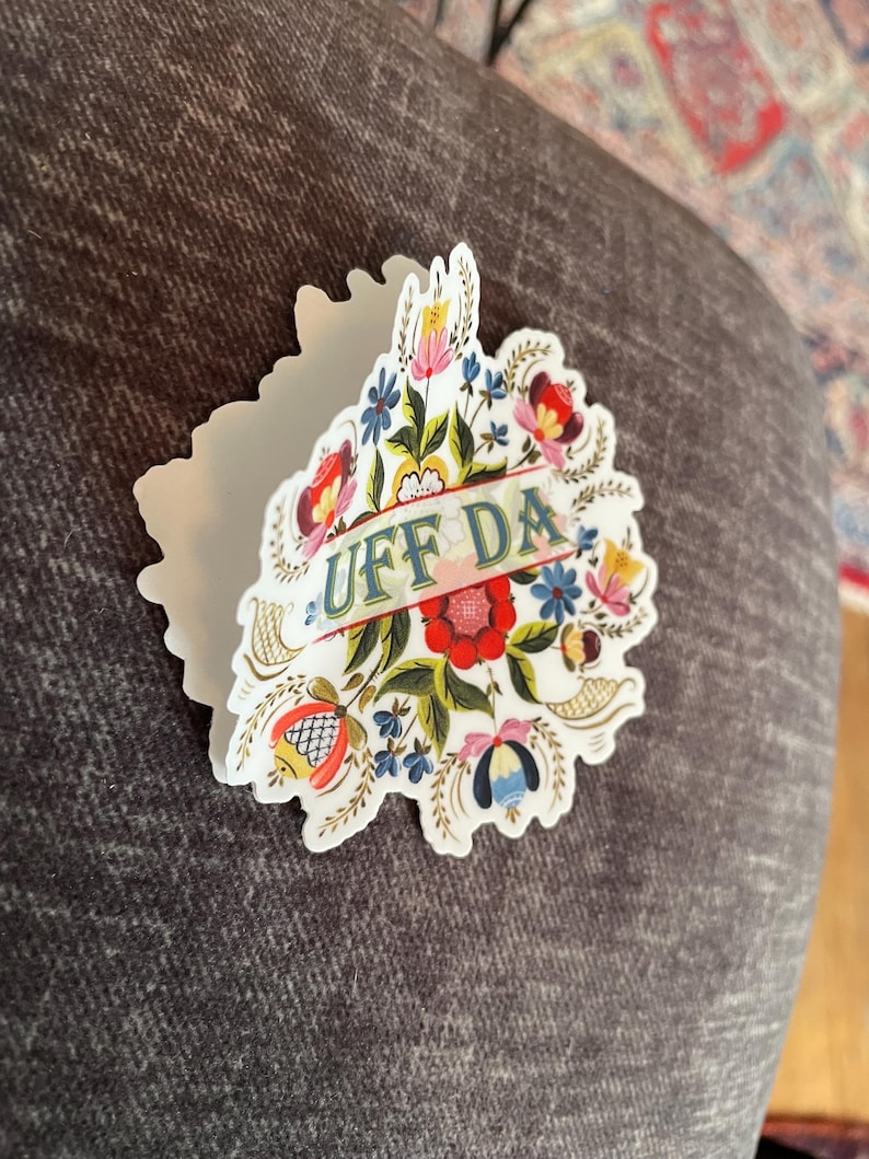 diecut adhesive sticker features colorful Norwegian rosemaling with the phrase ‘Uff Da’ written across the center