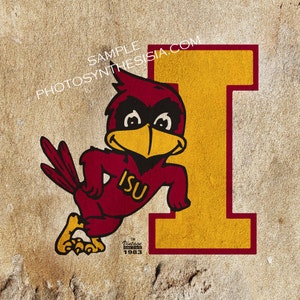 May include: A vintage 1983 Iowa State University logo featuring a red cardinal mascot with a yellow beak and a yellow letter "I" in the background. The cardinal is holding its right wing up and has the letters "ISU" on its chest.
